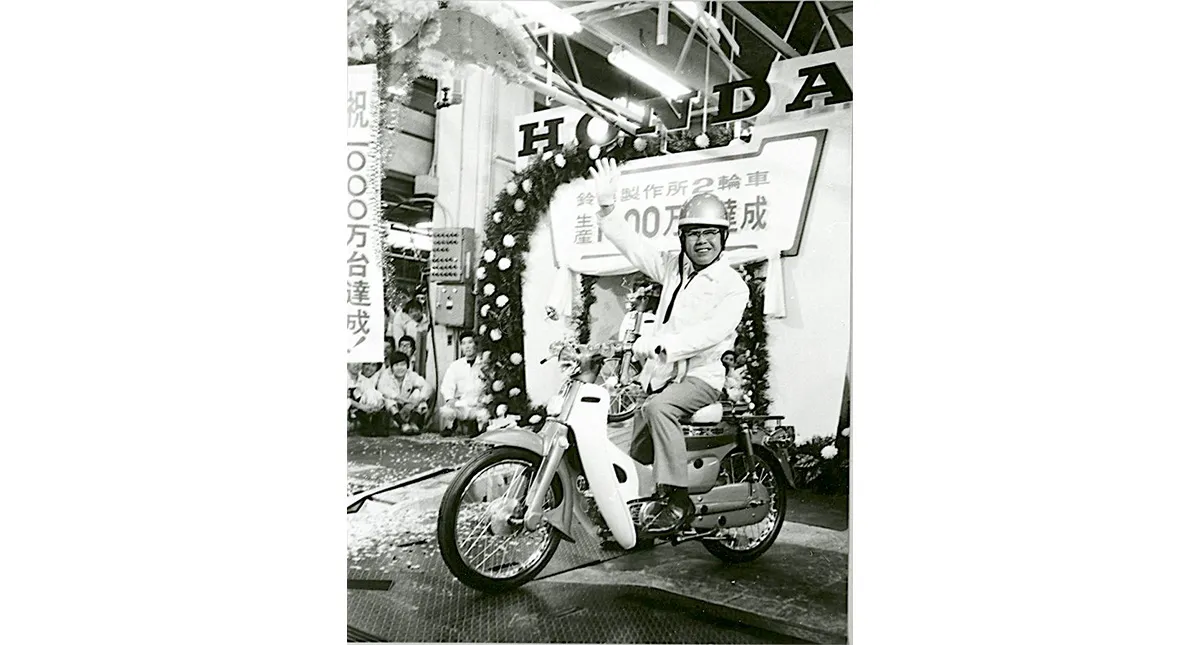 Soichiro Honda sorrindo e acenando enquanto está sentado em uma motocicleta Honda Super Cub durante uma celebração em uma fábrica.