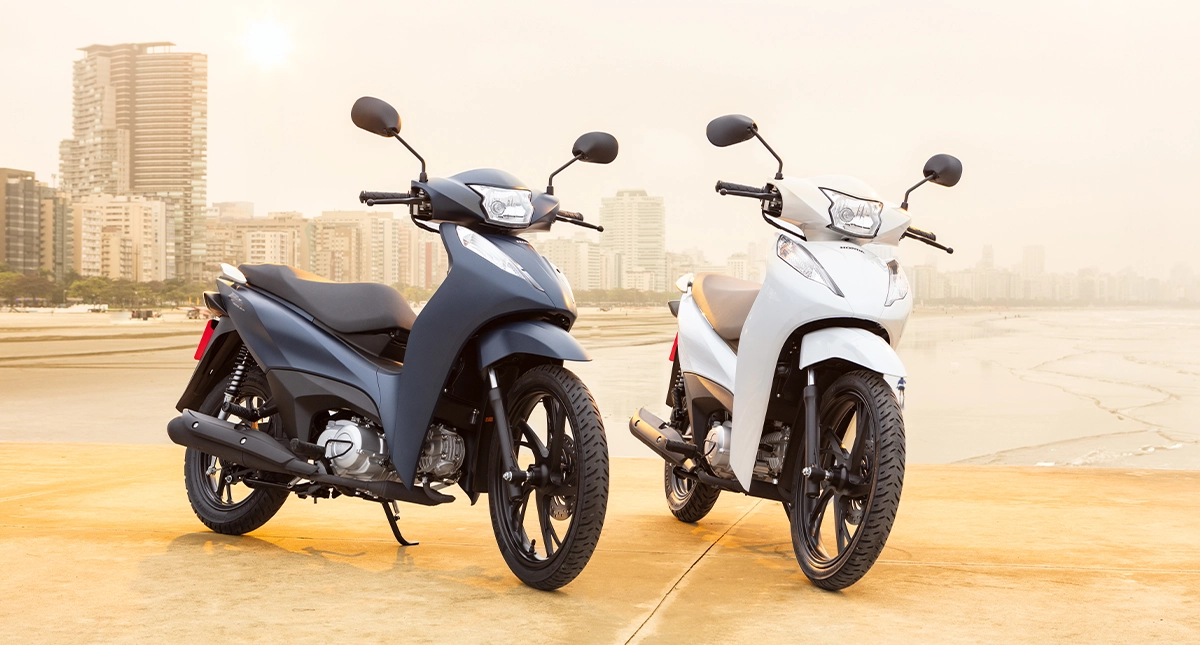 Duas motocicletas honda biz 125, nas cores azul marinho e branca, estcionadas na praia a beira mar