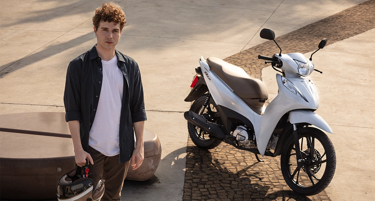 Homem com capacete em mãos ao lado da motocicleta honda biz 125 branca