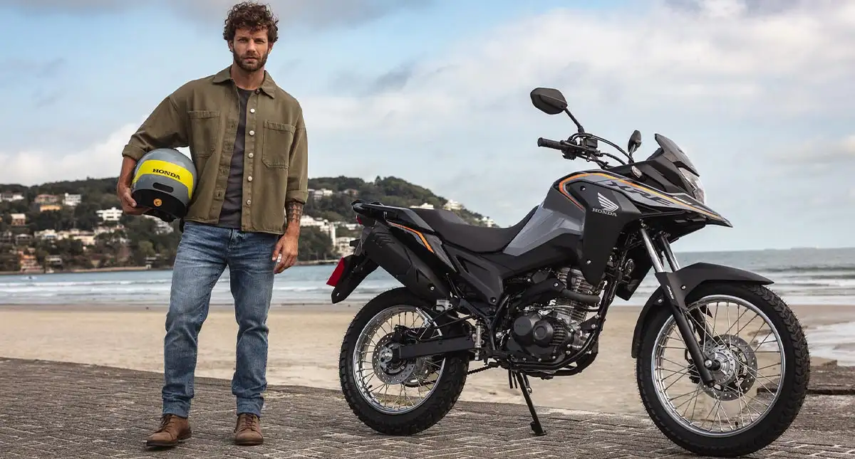 Homem com capacete na mão ao lado da motocicleta XRE 190 na praia