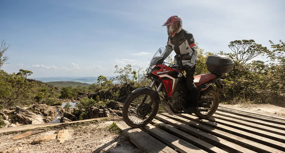 Piloto em pé na sua moto Vermelha Honda Sahara 300 passando por ponte de madeira