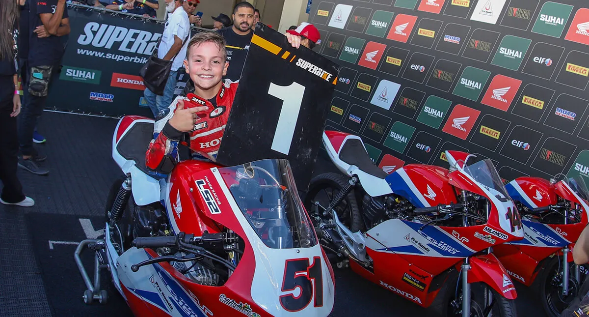 Piloto Enzo Laranjeira em um estande segurando uma placa com número um em mãos ao lado de três motocicletas Honda