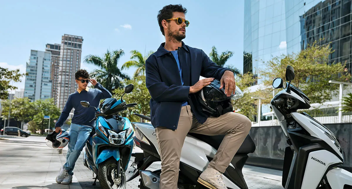 Em cenário urbano, dois homens estão encostados em suas Motocicletas Honda Elite nas cores Azul Metálico - Mystery Blue Metallic e Branco - Rosse White