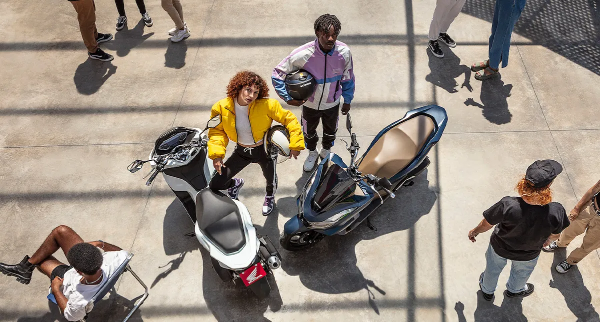 Homem e mulher com seus capacetes em mãos ao lado de suas Honda PCX em meio a outras pessoas 