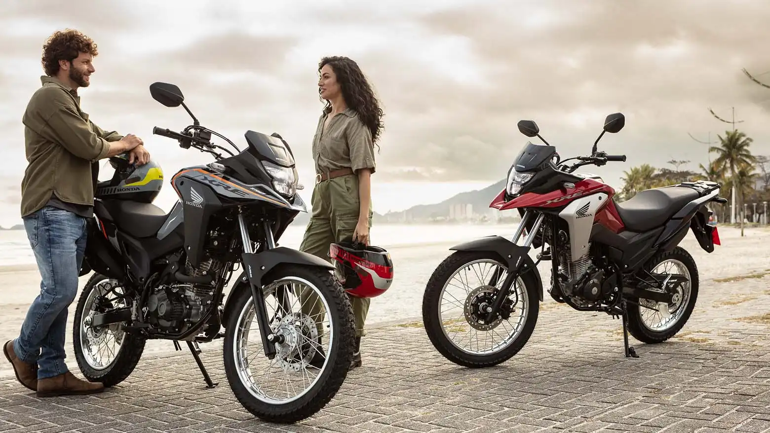Homem e mulher se olhando ao lado das Motos Honda XRE 190 com praia ao fundo 