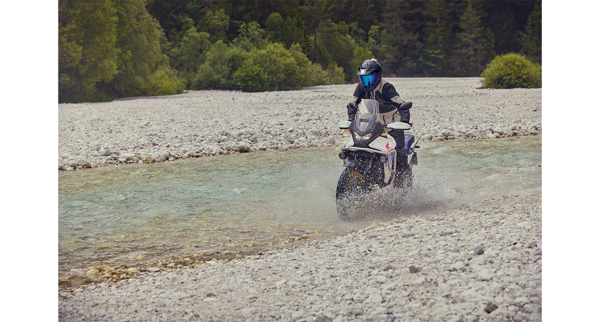 Piloto numa Honda Transalp em um pequeno rio com pedras ao lado 